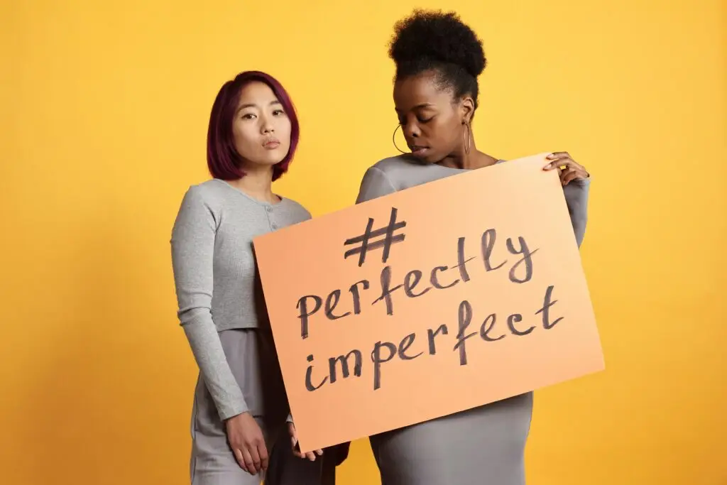 Two diverse women holding a sign that reads 'perfectly imperfect' against a vibrant yellow background.