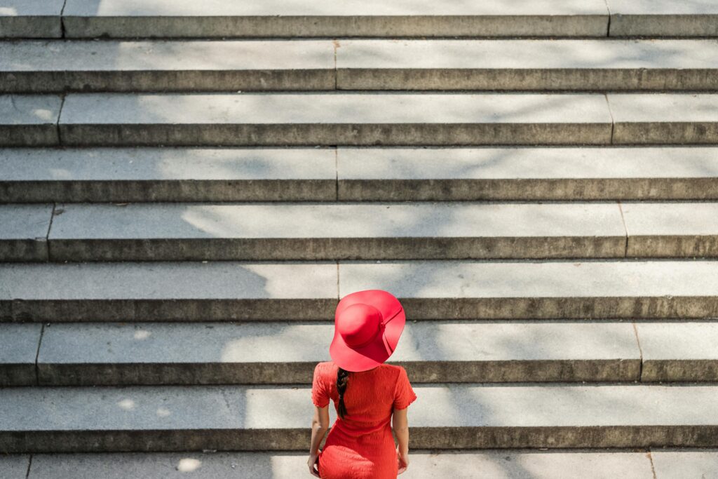 Woman in red dress and hat climbing shadow-dappled stairs outdoors.