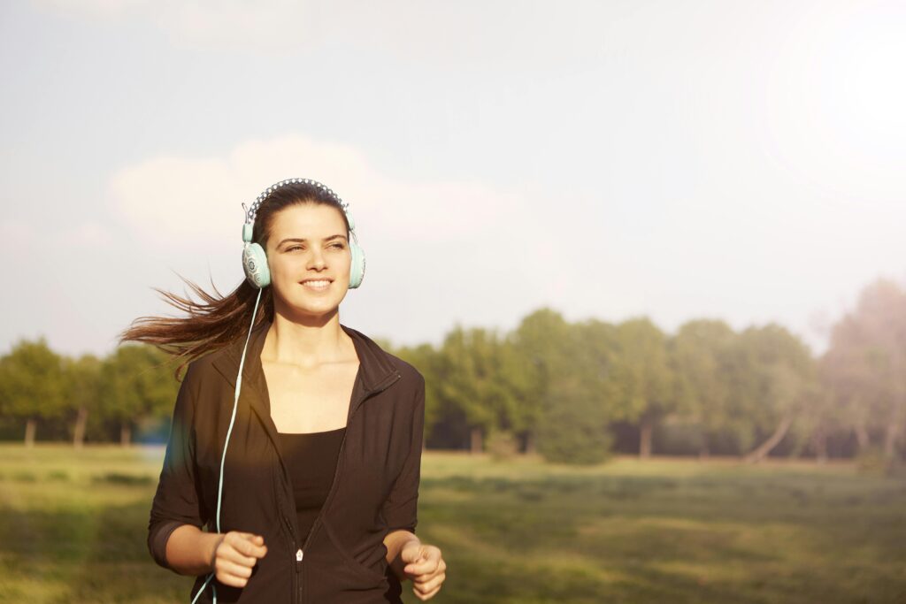 Smiling woman jogging in a park, listening to music with headphones, and enjoying the sunny day.